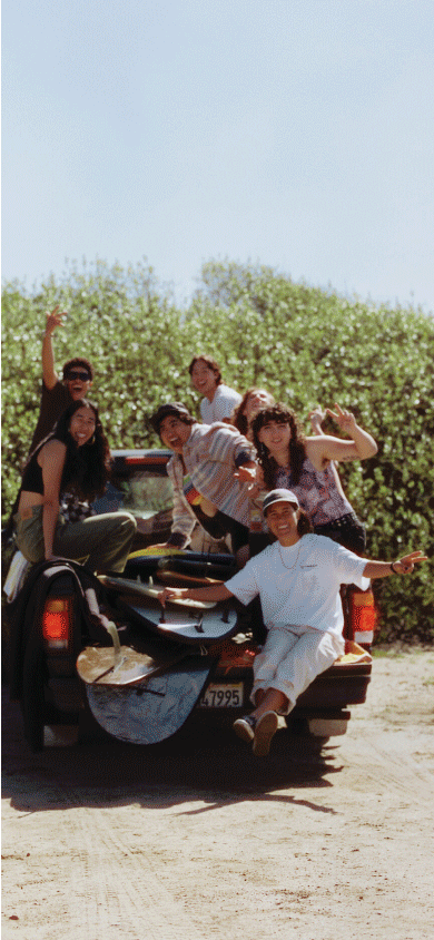 A group of friends sit in the back of a truck with their surfboards and smile for the camera in Vans clothes.