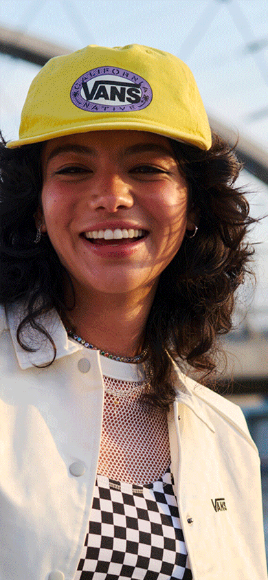A young woman smiles wearing a Checkerboard mesh top and a yellow Vans hat with a bridge in the background to create an urban scene.