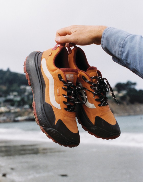 A hand holding up a pair of orange MTE Crosspath shoes with a white SideStripe.