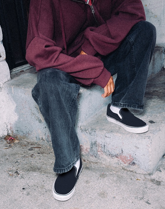 A person sitting on cement stairs wearing black slip on Vans shoes and a burgundy zip jacket