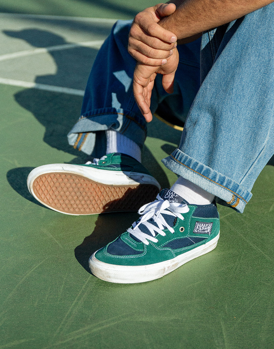 A mans sitting on the concrete wearing blue and green Half Cab styled with blue jeans.