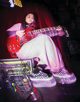 The Voice of Baceprot member is in casual clothes, wearing black and white checkerboard Vans Old Skool shoes sitting on an amplifier and playing an electric guitar on stage.