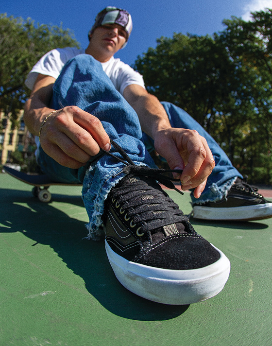 Skateboarder Etienne Gagné sitting on a skateboard, tying Skate Old Skool 36+ shoes on a bright green surface.Skateboarder Etienne Gagné sitting on a skateboard, tying Skate Old Skool 36+ shoes on a bright green surface.