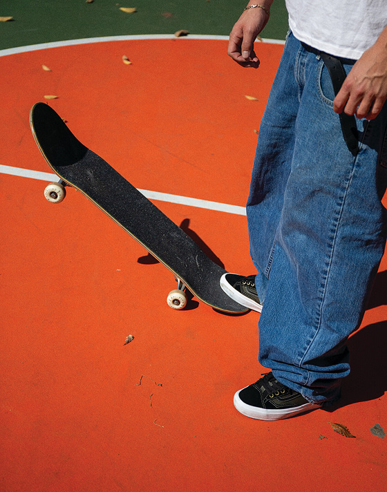A person in baggy jeans stands on a bright orange floor, resting a skateboard on one foot and wearing Skate Old Skool 36+ shoes.