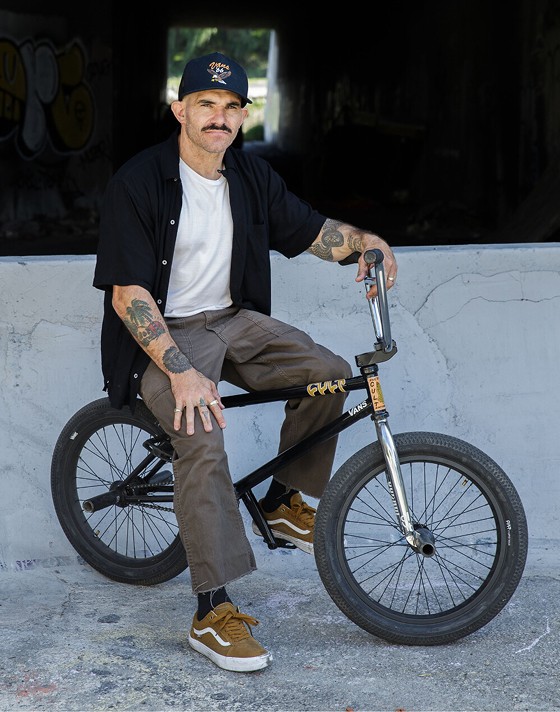 A man with tattoos and a mustache is sitting on a BMX bike while wearing Golden Brown BMX Old Skool shoes in front of a tunnel. 