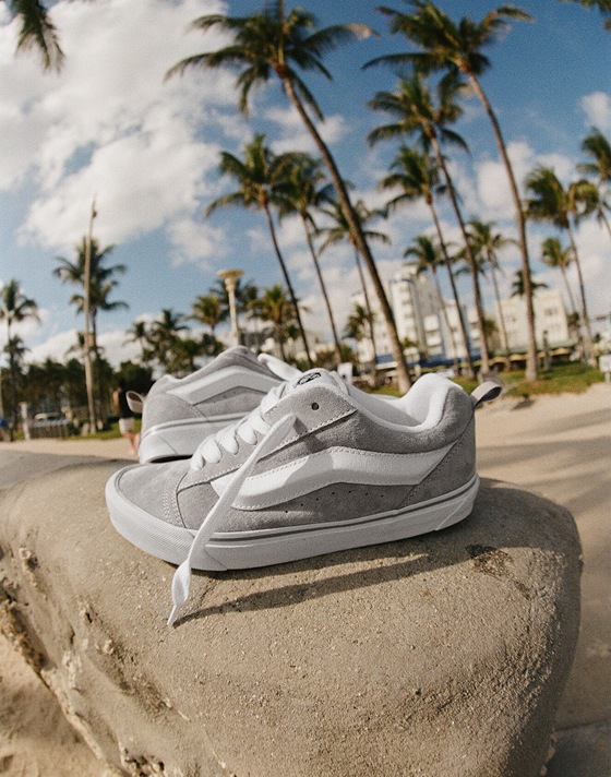 A pair of Pig Suede Grey Knu Skool shoes rests on a stone wall. Palm trees and a vibrant blue sky are visible in the background.