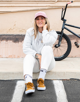 A smiling woman in a pink cap,gray sweater and Golden Brown BMX Old Skool shoes sits casually a curb next to a BMX bike