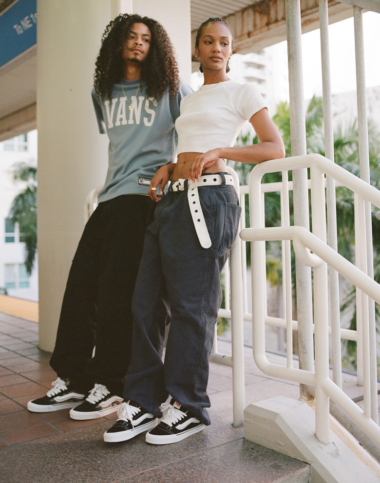 A man and woman lean against a staircase railing, both wearing Vans apparel and looking away from the camera. 