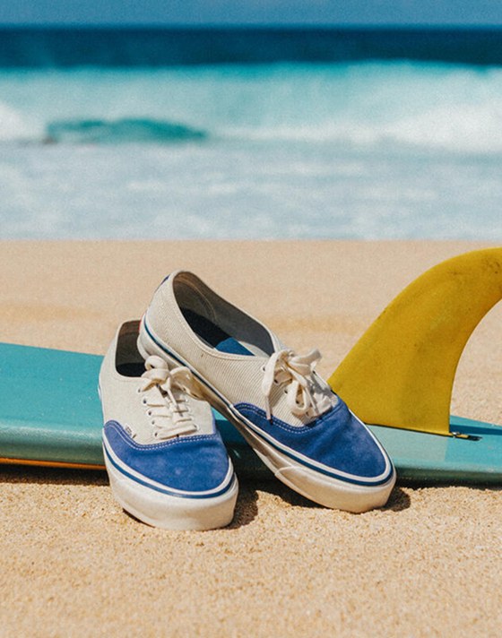 A pair of Authentic Reissue sneakers in Thalia Blue sit on a multicolored surfboard lying in the sand. The ocean can be seen out of focus in the background.