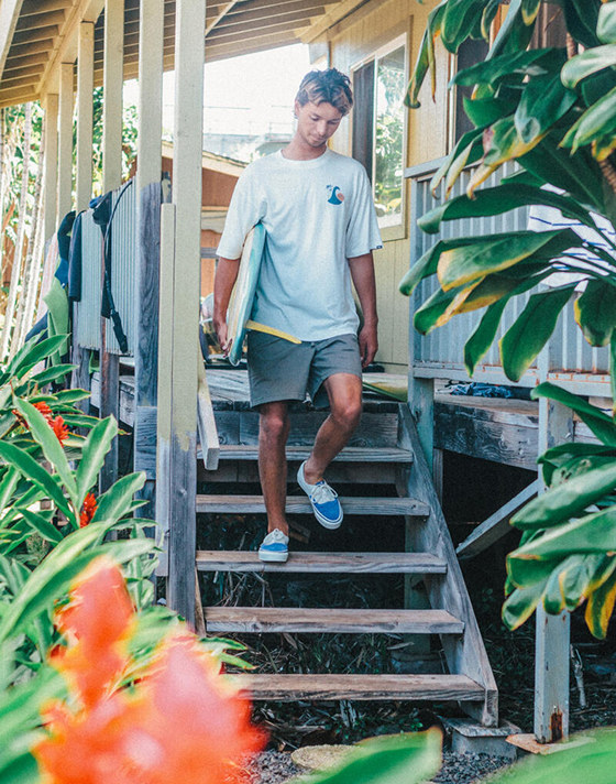 Surfer Tosh Tudor is walking down wooden stairs of a deck while holding a blue and white surfboard. Tudor wears a white t-shirt, shorts, and Authentic Reissue sneakers in Thalia Blue.