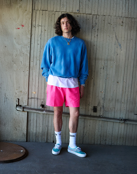 A person sitting on the ground infront of a graffiti wall, wearing a white Vans crew neck sweatshirt, white socks and white authentic shoes with blue gumsole.