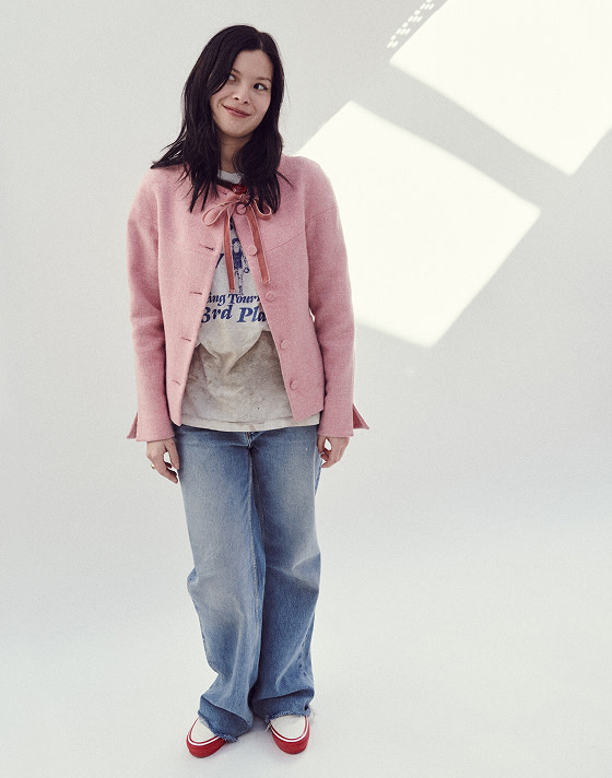 A person stands against a white studio background, facing the camera, with their arms at their sides. They are wearing a pink overcoat, a white t-shirt, washed blue jeans, and Premium Authentic 44 sneakers in Duck Canvas White & Red.