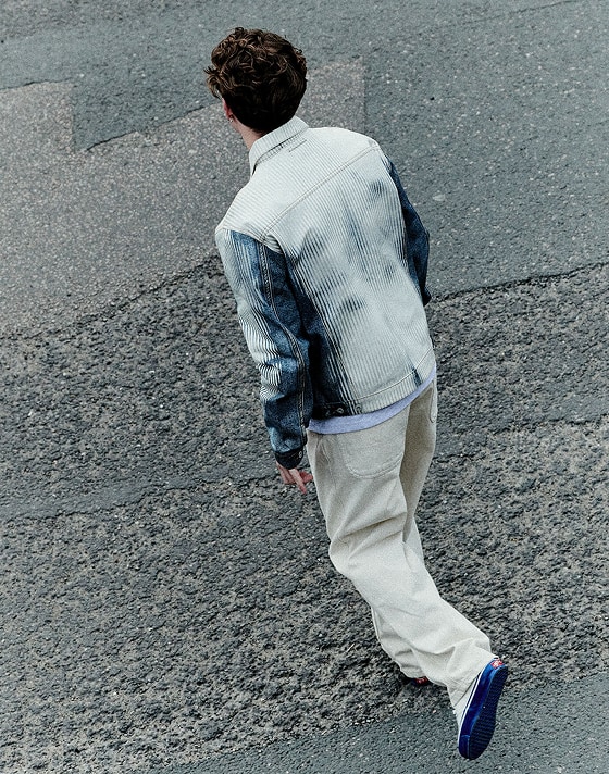 A Dutch angle shot of person walking away from the camera across a road. They are wearing a white and blue denim jacket, cream jeans, and Premium Authentic 44 sneakers in Duck Canvas White & Blue.