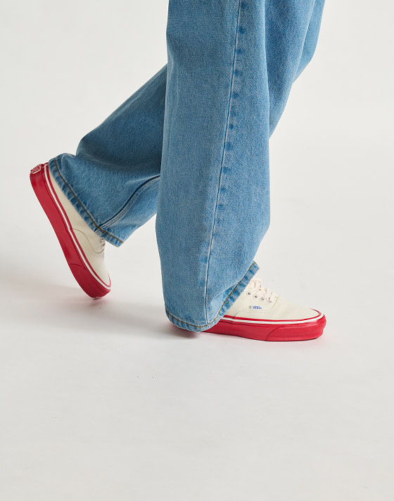 A detail knee-down shot of a person walking to the right against a white studio background. They are wearing washed blue jeans and Premium Authentic 44 sneakers in Duck Canvas White & Red.