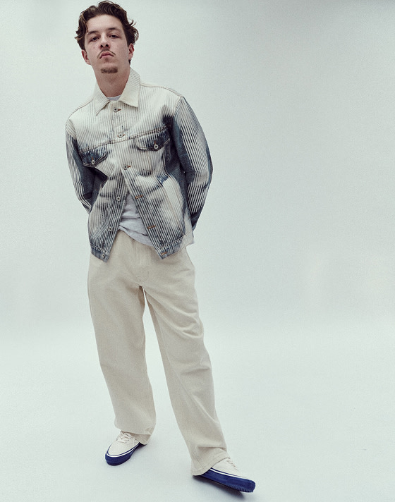A person stands against a white studio background, facing the camera, with their arms behind their back. They are wearing a white and blue denim jacket, cream jeans, and Premium Authentic 44 sneakers in Duck Canvas White & Blue.