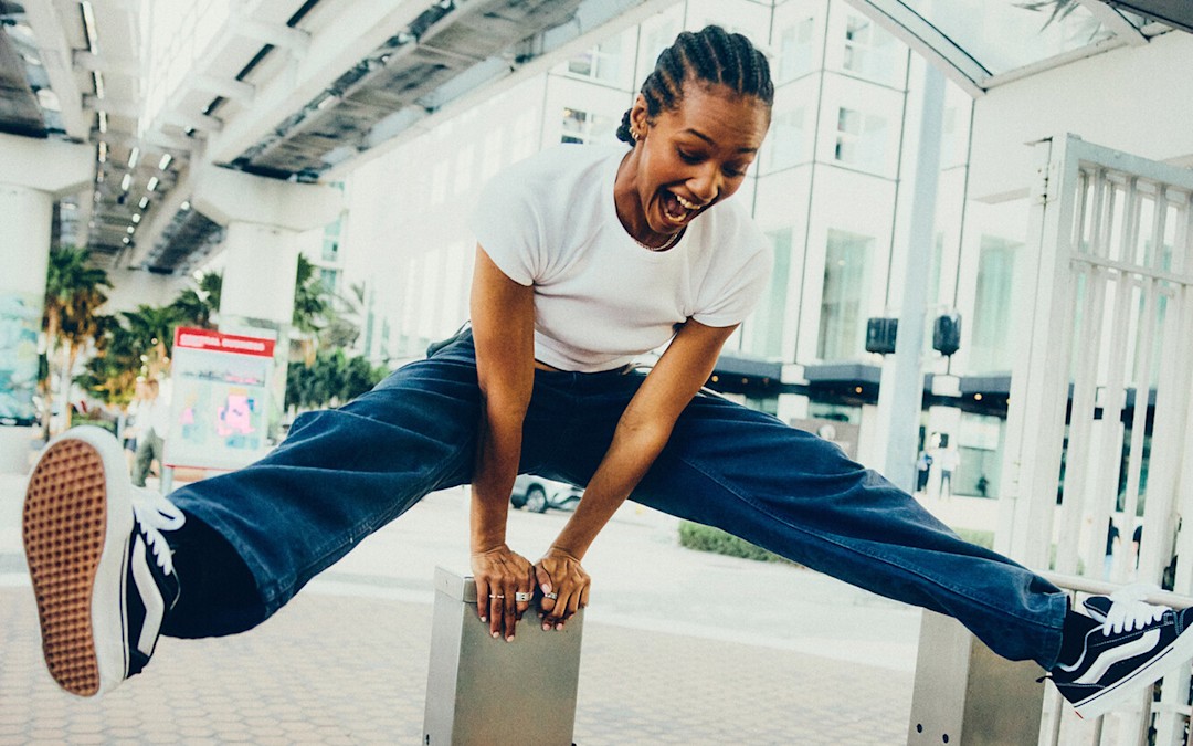 A person jumps over a post in a train station with their legs outstretched to both sides. They are wearing a white t shirt, blue jeans, and black and white Knu Skool shoes.