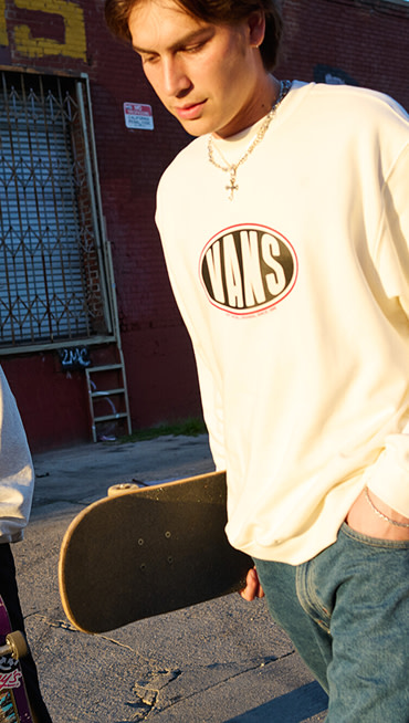 A man walks down a street in a Vans long-sleeve shirt holding a skateboard at golden hour.