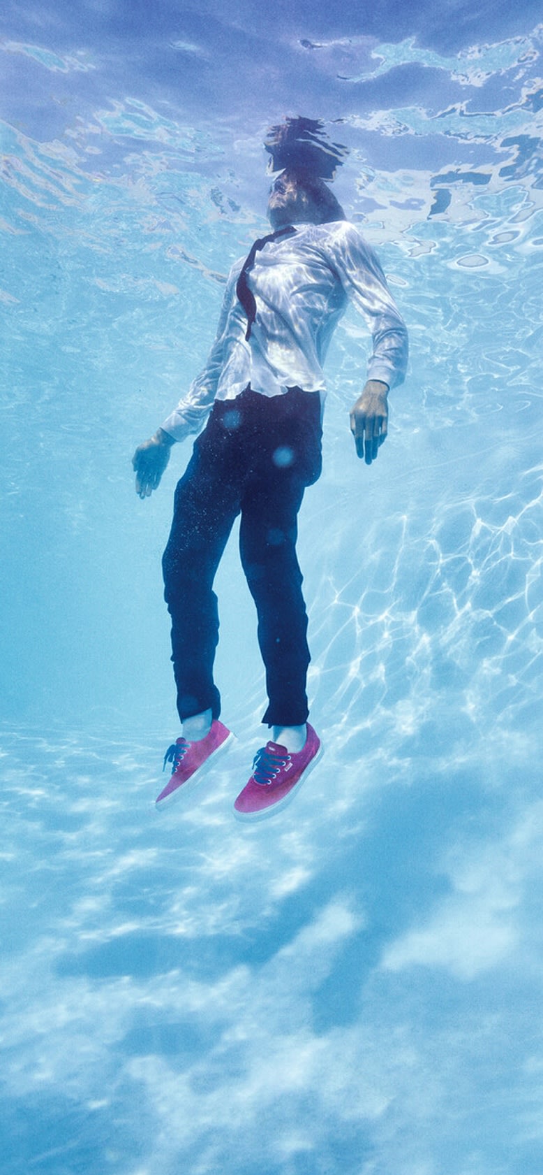 An underwater shot of Vans skater Curren Caples floating in a pool in a white collared shirt, black tie, black pants, and red Curren shoes.