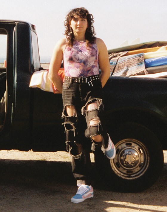 A person poses in front of a truck full of surfboards wearing ripped black jeans and blue and pink Knu Skool.
