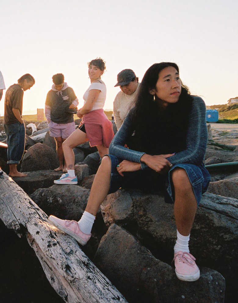 A group of friends hang out and talk on some rocks at the beach during golden hour, wearing Vans clothes and shoes.
