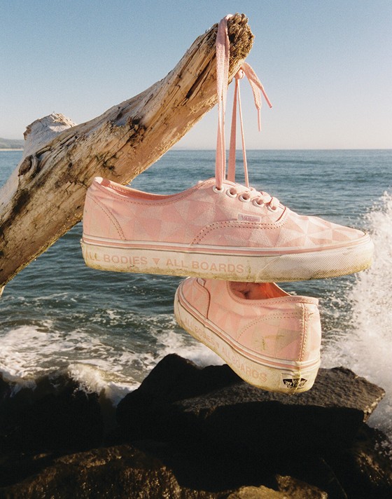 A pair of pink Authentic hang on a piece of driftwood by their laces that say "All Bodies, All Boards" on the rubber sidewalls.