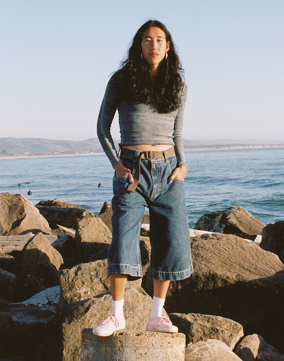 Someone poses for the camera wearing pink Authentic and baggy denim capri pants with the ocean behind them.