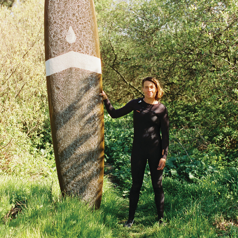 A surfer holds their longboard upright in a wetsuit surround by foliage near a beach.