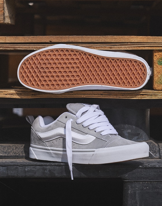 A gray and white Knu Skool shoe rests on a wooden surface as another is wedged in between a wooden pallet so the sole is visible.