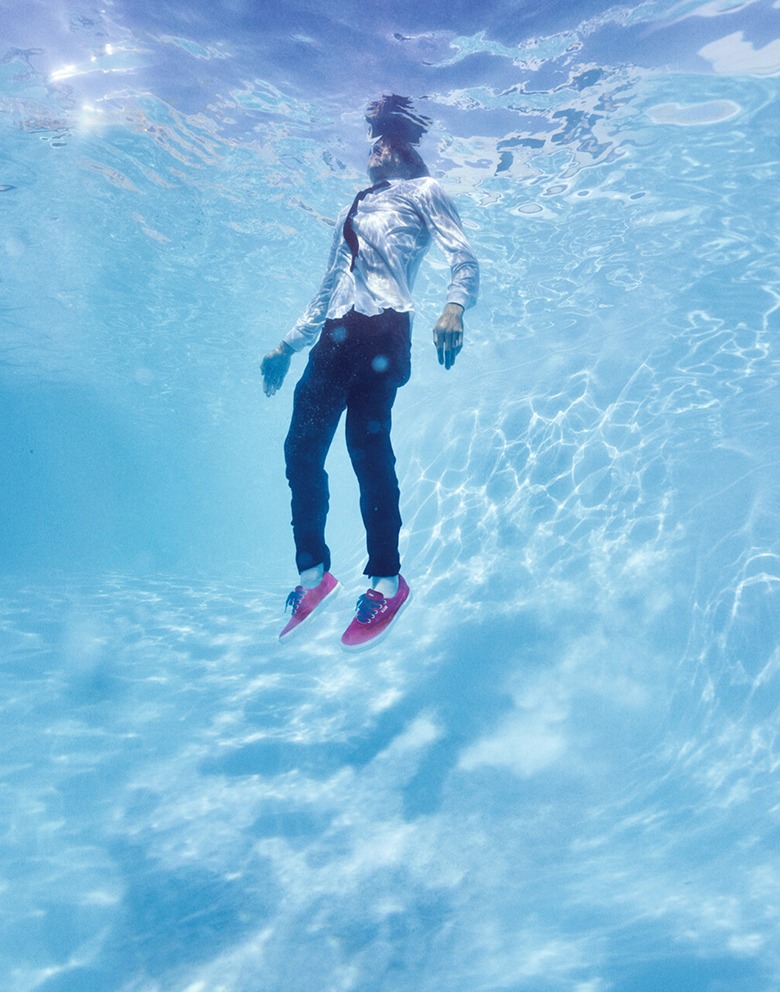 An underwater shot of Vans skater Curren Caples floating in a pool in a white collared shirt, black tie, black pants, and red Curren shoes.