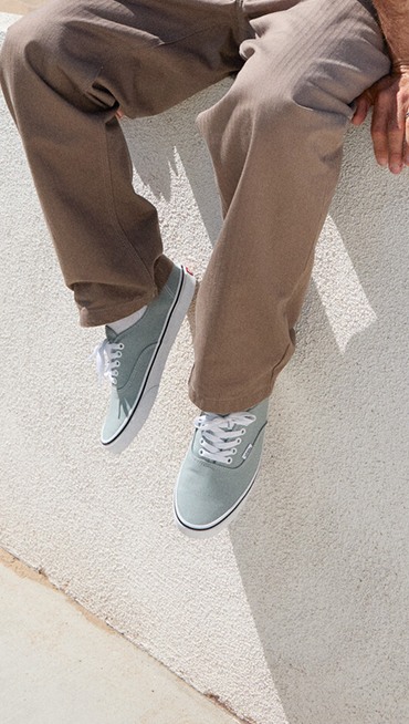 Close-up of a man sitting on a white stucco wall wearing Pure Grey Authentic and brown pants.