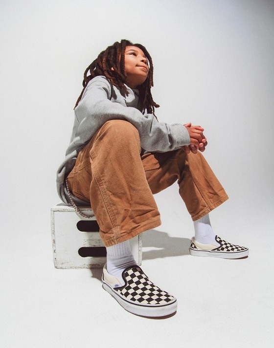 A boy sits on a wooden box wearing a gray hoodie, brown jeans, and black and white Checkerboard Slip-On.