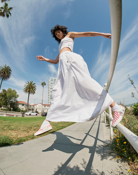 A woman jumps from a rail looking into the camera wearing a white crop top, a white dress, and Honeysuckle Pink Checkerboard Classic Slip-On.
