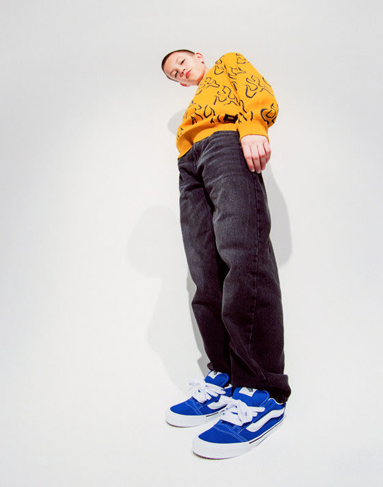 A boy looks down into the camera in a photography studio wearing a yellow sweater with black flames and blue Knu Skool shoes.