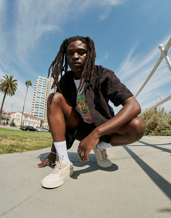 A man squats and looks into the camera on a concrete walkway wearing Vans clothes and Warm Beige Old Skool.