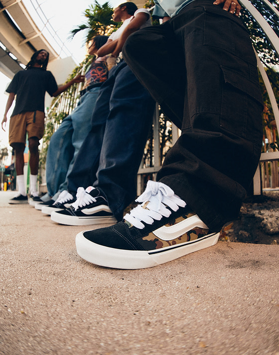 A close-up of a pair of camo Knu Skool on a man wearing black jeans with a group of young people in the background under a tram line.