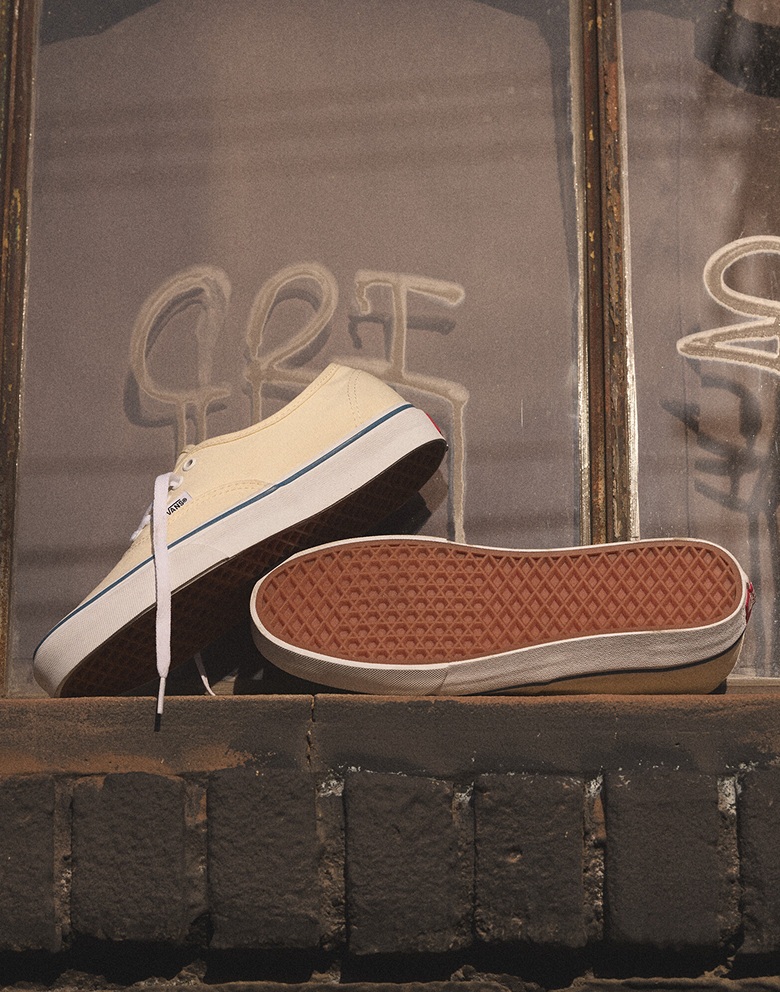 A pair of white and blue Premium Authentic rests on a windowsill stacked, one shoe showing its sole to the camera, with graffiti in the background.