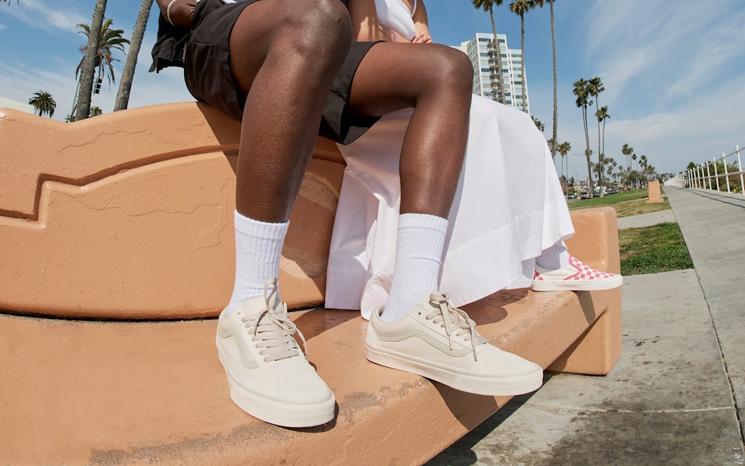 A close-up of two people wearing Warm Beige Old Skool and Honeysuckle Pink Checkerboard Classic Slip-On on a concrete bench.