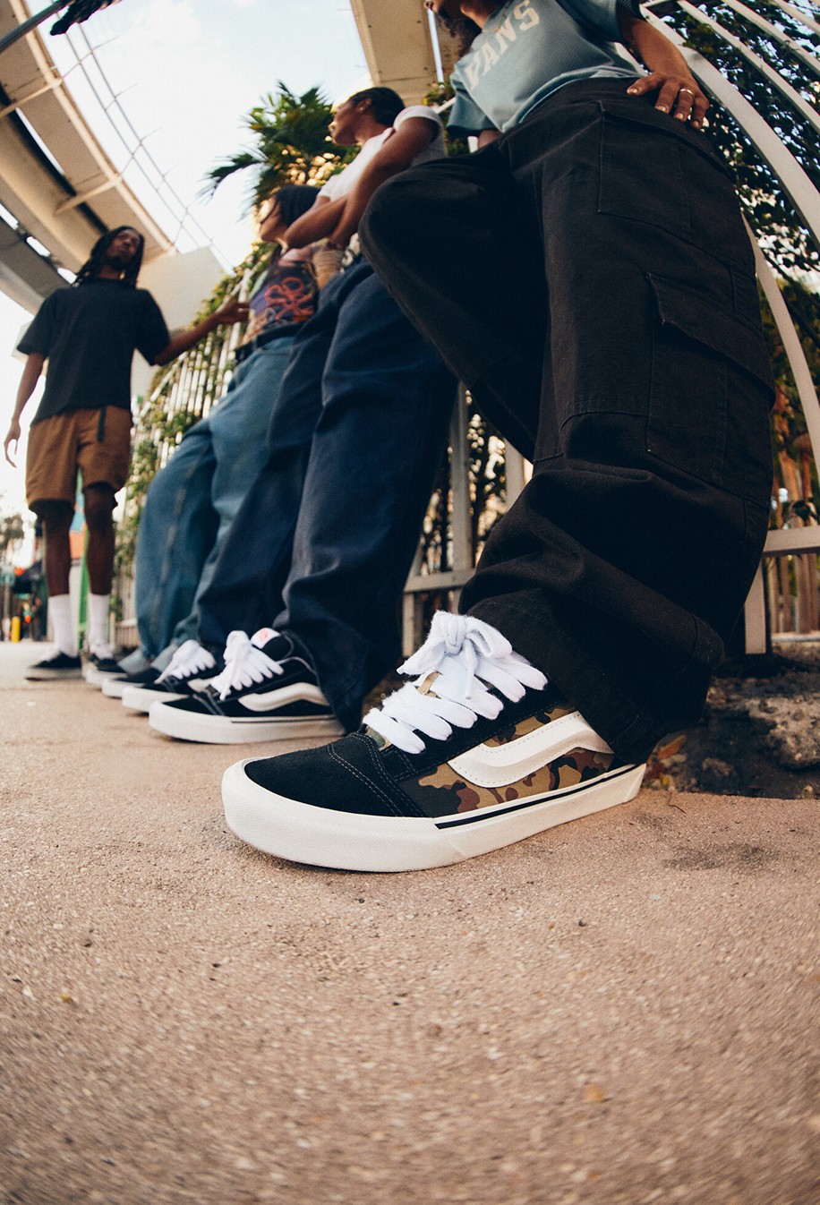 A close-up of a pair of camo Knu Skool on a man wearing black jeans with a group of young people in the background under a tram line.