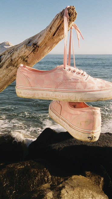 A pair of Coral Brush Pink Vans X Queer Surf Authentic shoes hang from a piece of driftwood by their laces with waves breaking in the background.