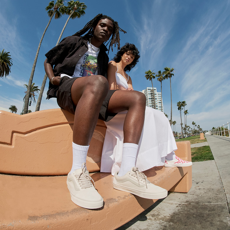 Two people wearing Warm Beige Old Skool and Honeysuckle Pink Checkerboard Classic Slip-On on a concrete bench.