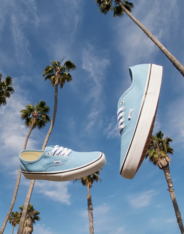 A pair of Heritage Blue Vans Authentic Shoes fly through the air as if thrown beneath palm trees and a blue sky with few clouds.