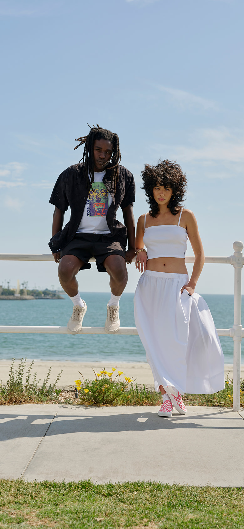 A man sits on a railing wearing Warm Beige Old Skool and black clothes at the beach. The woman next to him wears a white crop top and dress.