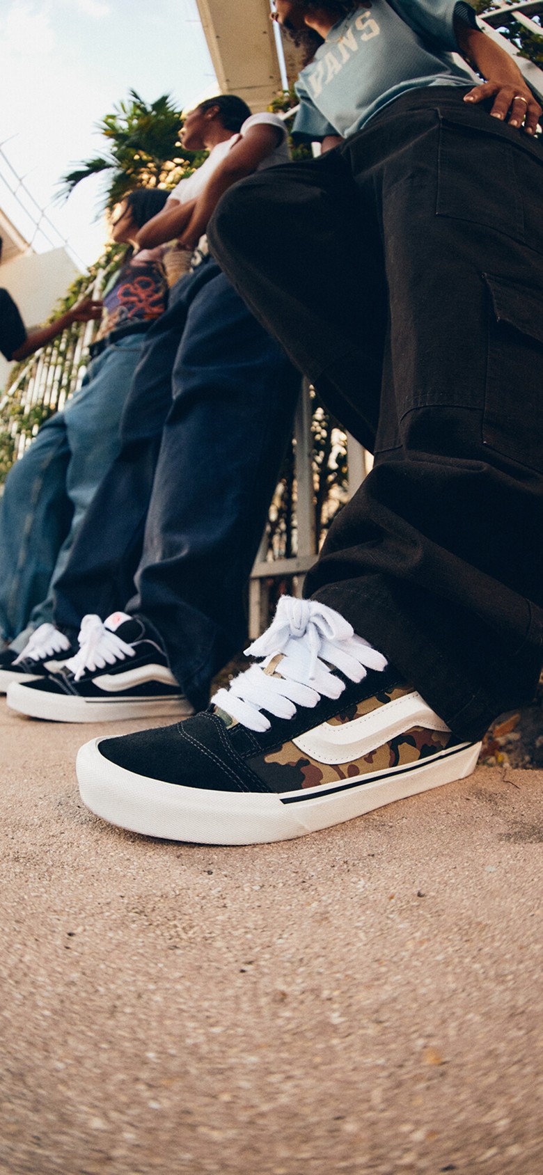 A close-up of a pair of camo Knu Skool on a man wearing black jeans with a group of young people in the background under a tram line.