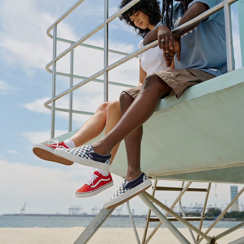 Two friends sit on a lifeguard tower kicing their red Old Skool and blue Checkerboard Classic Slip-On in the air with the beach behind them.
