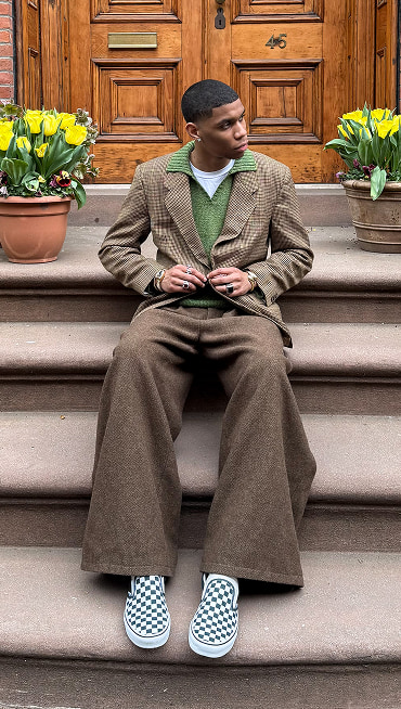 @nassnyc sits on a staircase wearing a brown houndstooth blazer, baggy tweed pants, and white and black Checkerboard Classic Slip-On.