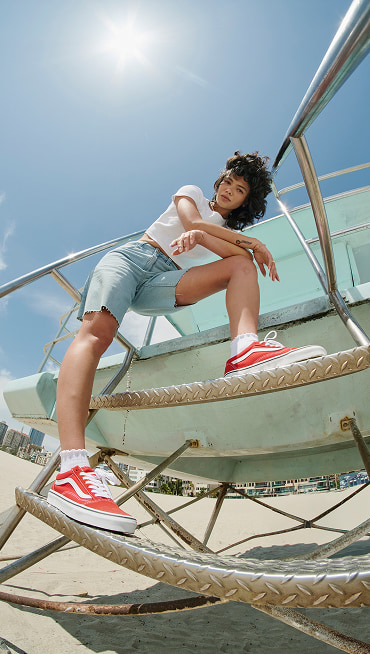 A young woman stands on the steps of a lifeguard tower and looks into the camera wearing Racing Red Old Skool shoes with the sun high in the sky.