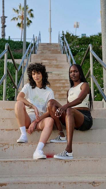 Two women sit on a staircase wearing white Authentic shoes and black and white Checkerboard Classic Slip-On near a beach.