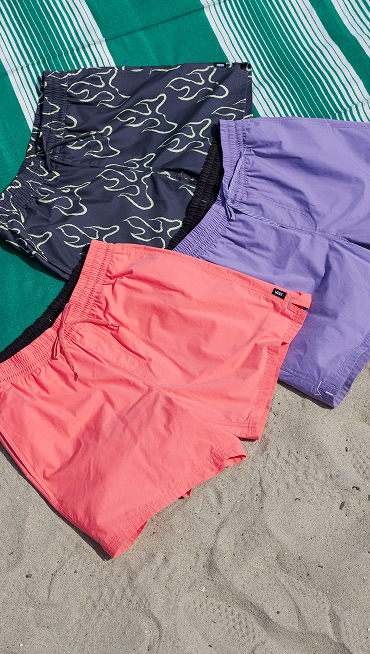 The Primary Solid Elastic 17'' Boardshorts in Porcelain Rose Pink, Purple Haze, and Asphalt Grey with Shadow Lime Green flames lay on the beach.