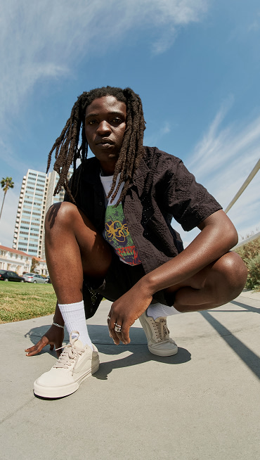 A young man squats on a sidewalk and looks into the camera wearing black Vans clothes and Warm Beige Old Skool.