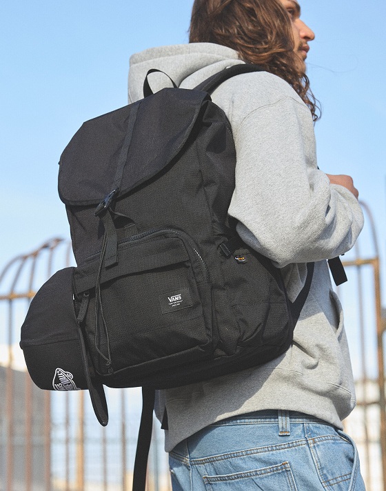 A man in a gray carries a black Startle Backpack slung over one shoulder.